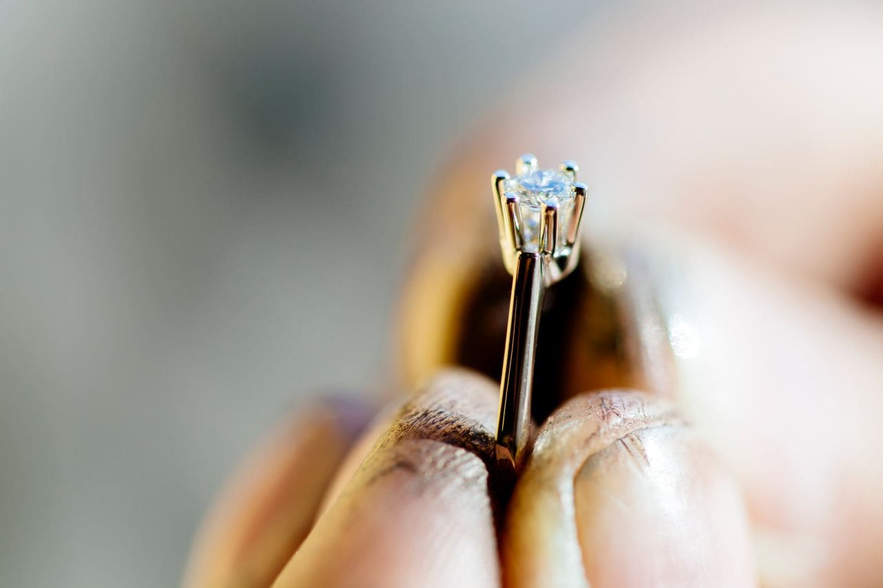 A jeweler with dirty hands holds a platinum solitaire ring with a prong-set, round cut moissanite center stone