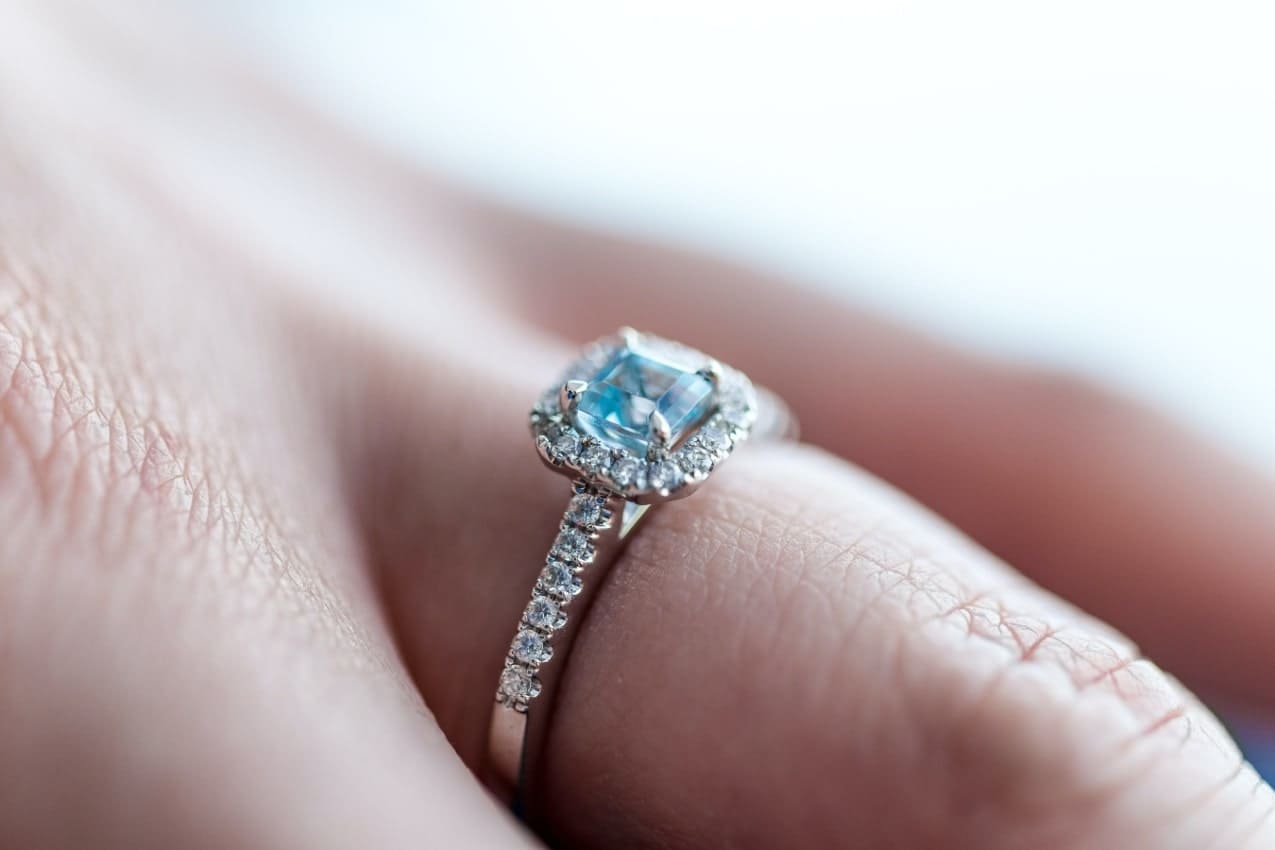 Close up image of a hand wearing a silver engagement ring with a blue, princess cut center stone