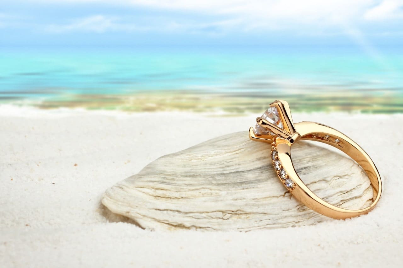 A gold, prong set ring leaning against an oyster shell on a beach