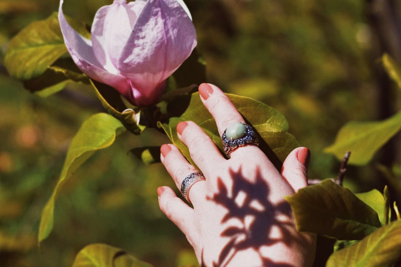 A woman’s hand wearing opal rings reaches toward a flower.