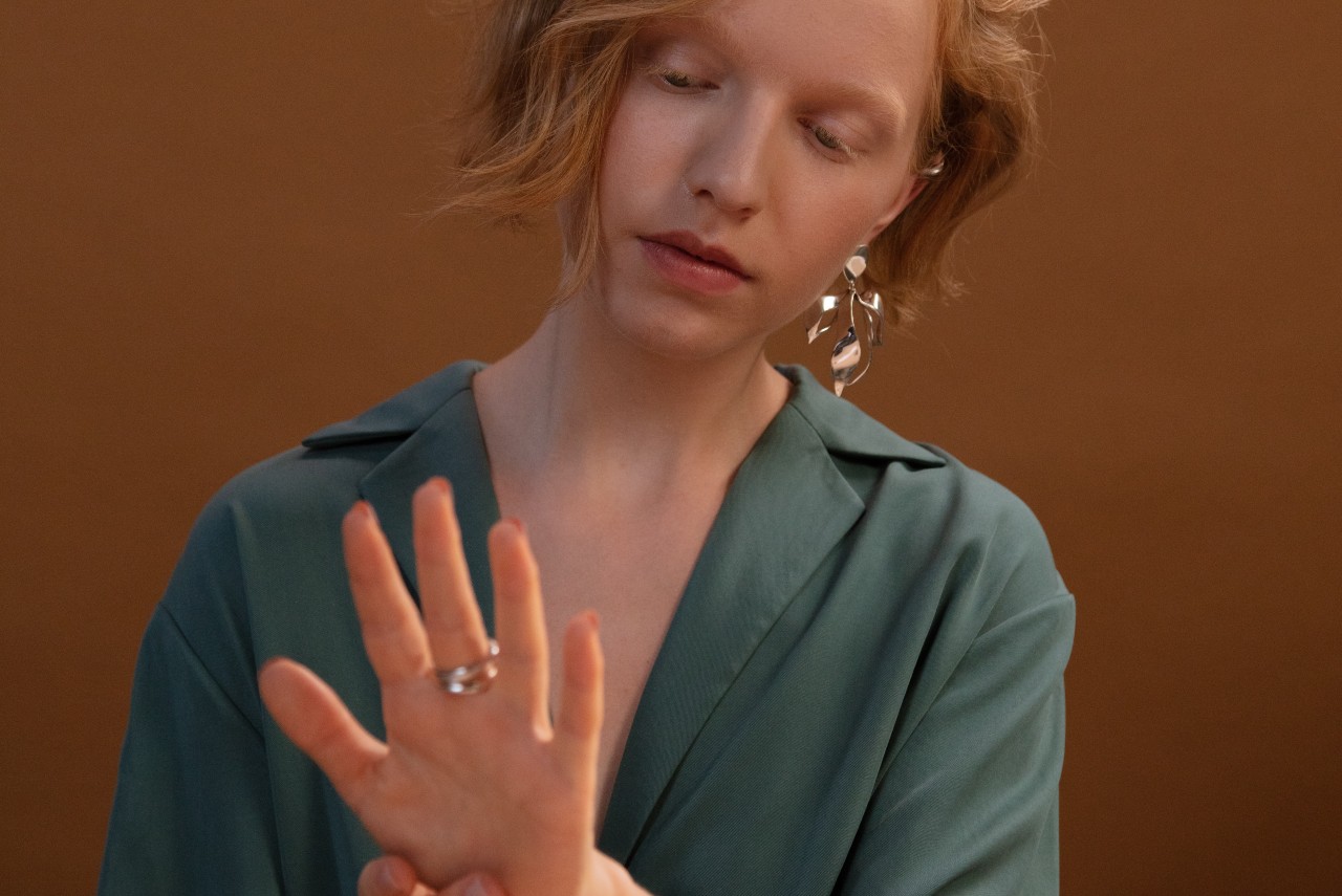 A red-haired woman inspects her white gold jewelry in front of a brown backdrop.