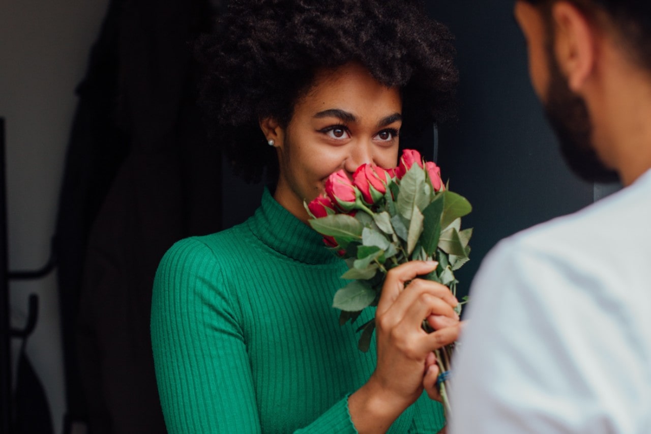 A woman wearing a green turtleneck smells roses while wearing pearl stud earrings.