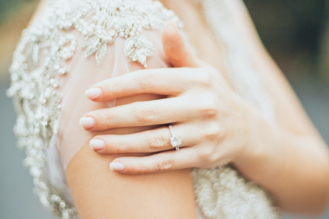 A bride touches her shoulder, wearing a side stone ring.