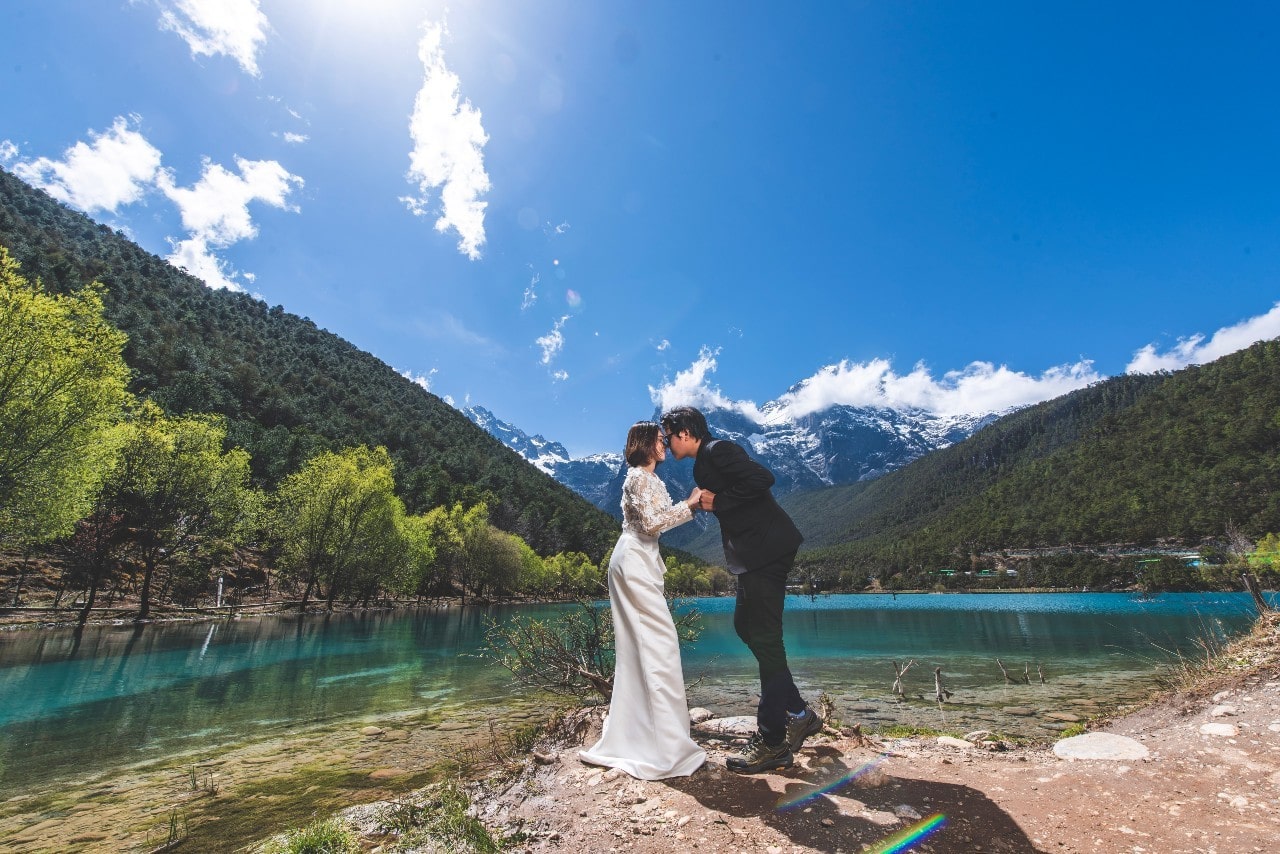 A bride and groom kiss by a pond in the mountains on a sunny day.